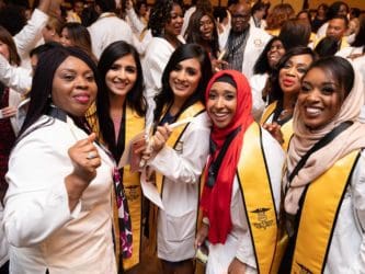 six women of color wearing white lab coats with honors at graduation