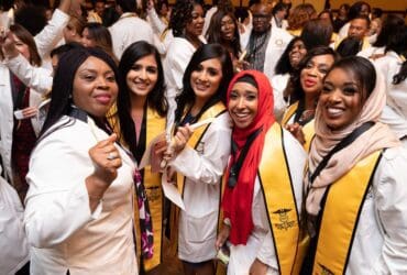 six women of color wearing white lab coats with honors at graduation